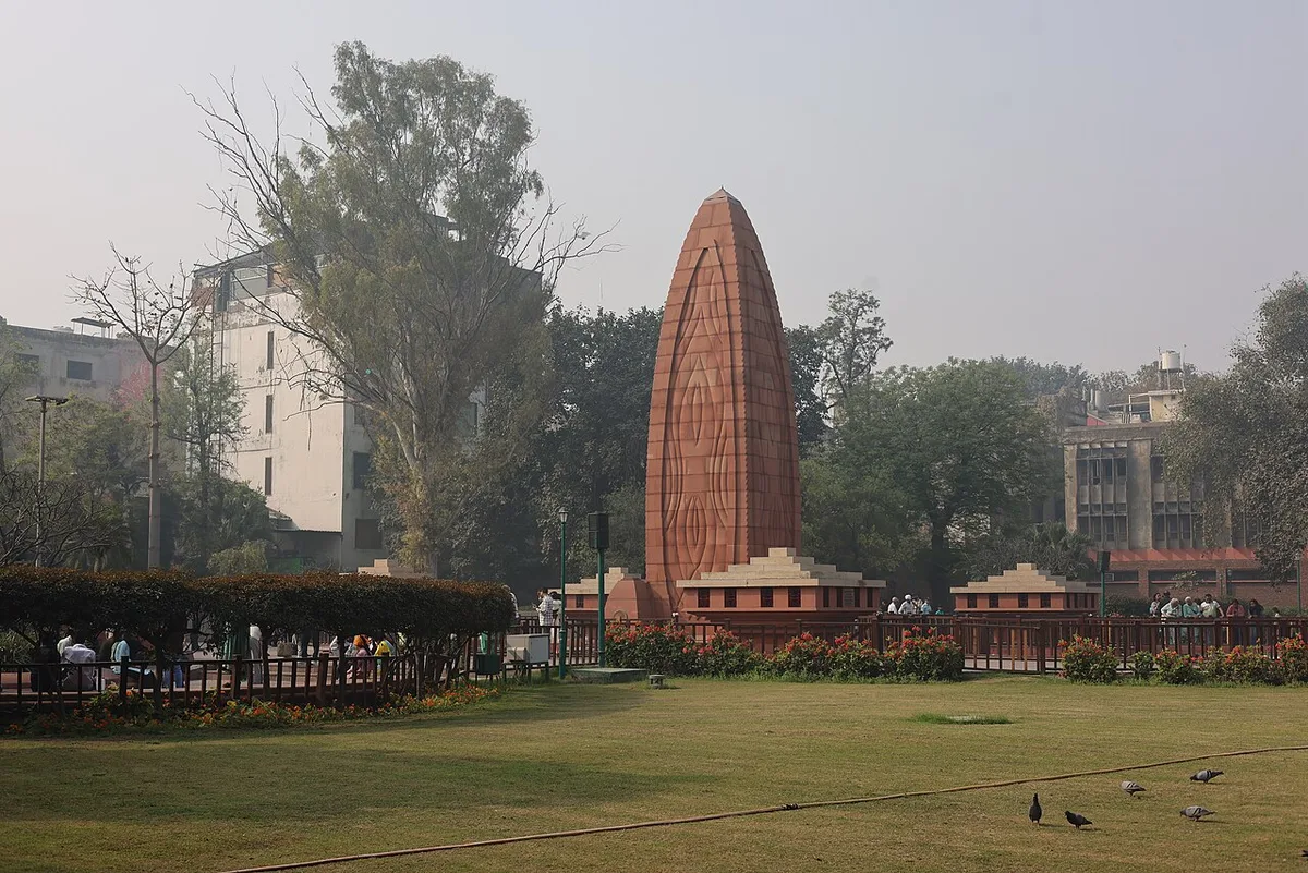 The flame-shaped red sandstone memorial tower at Jallianwala Bagh, standing over lawns and flower beds with visitors at its railing
