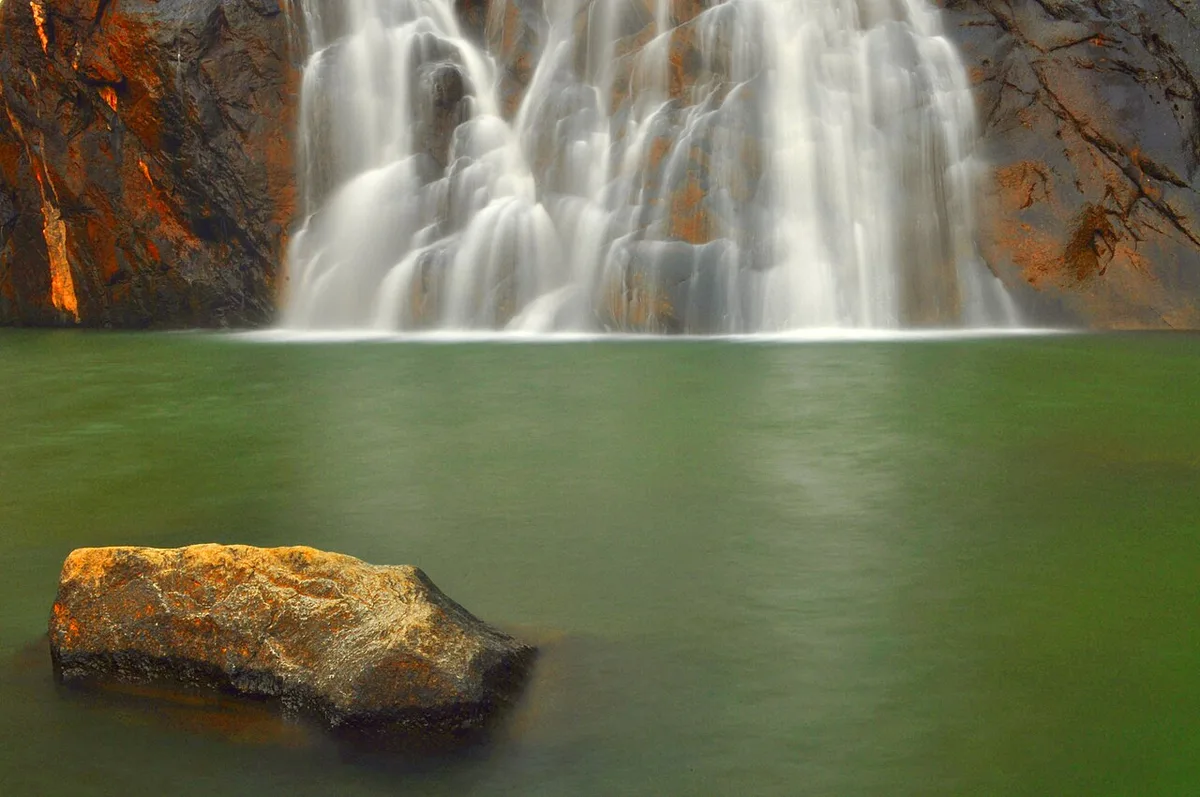 The lowest tier of Dudhsagar Falls pouring in white strands into a green pool, with a sunlit boulder in the foreground
