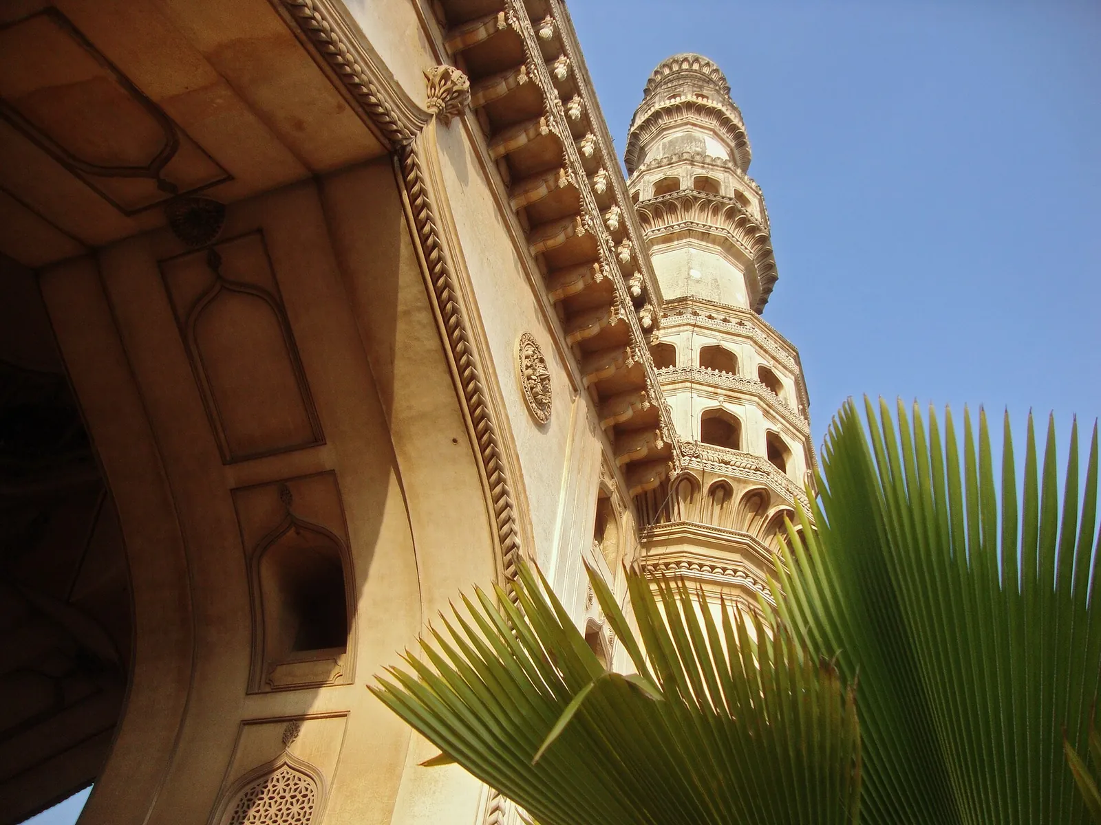 A minaret of the Charminar in Hyderabad against a clear blue sky, its carved balconies lit by the sun
