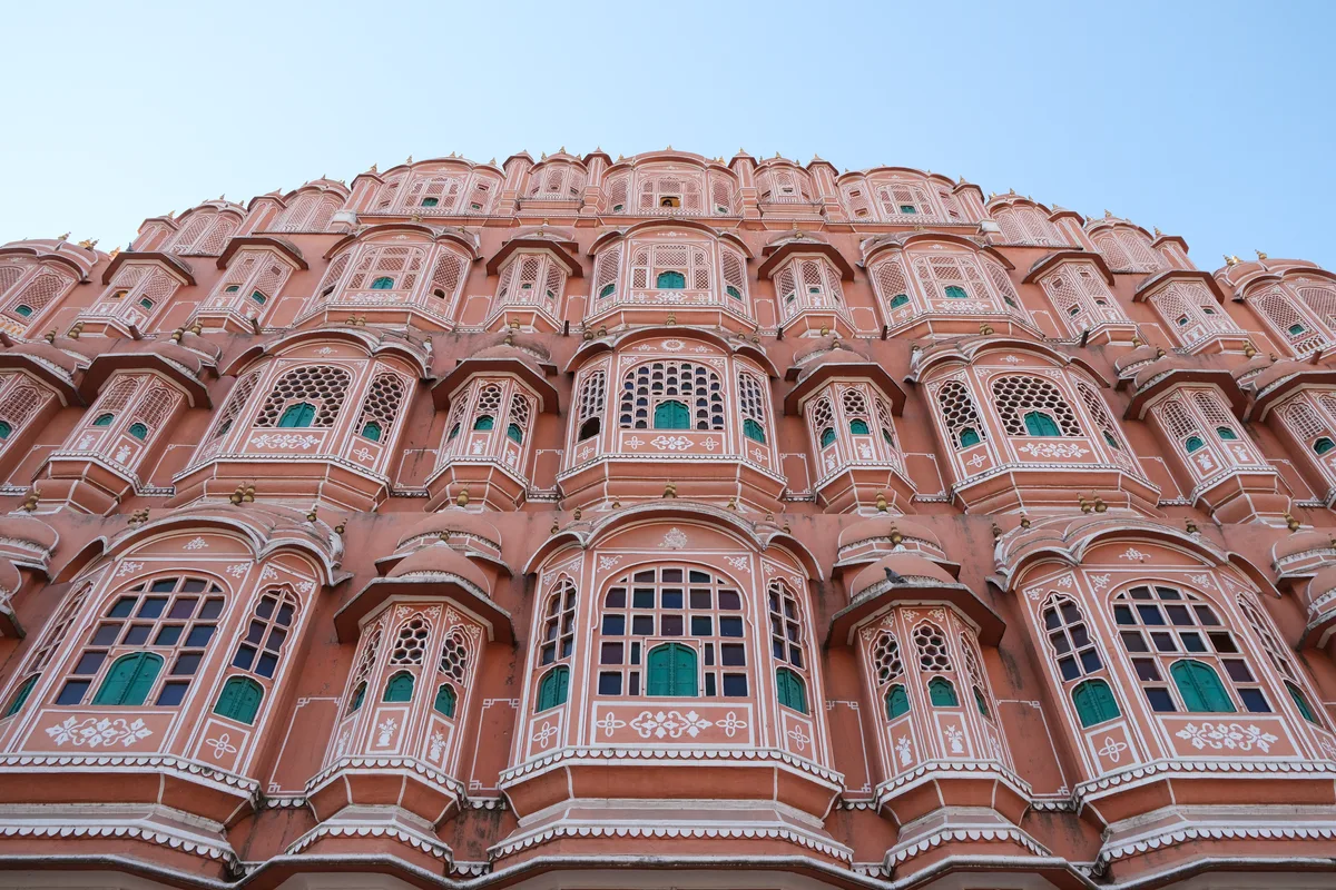 The pink sandstone facade of the Hawa Mahal in Jaipur, its tiers of latticed windows and small domed balconies seen from below against a clear sky