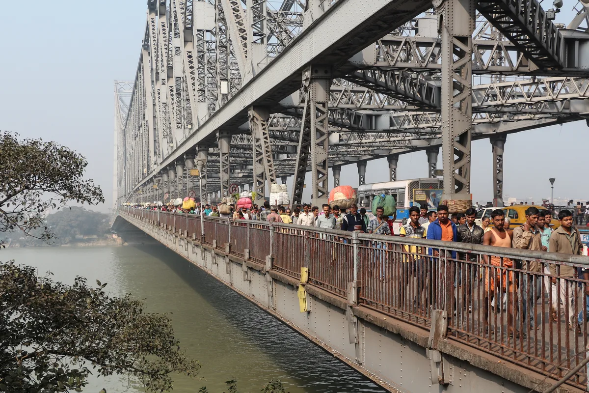Pedestrians crossing the Howrah Bridge on its footpath, the steel cantilever trusses rising above them and the Hooghly river below