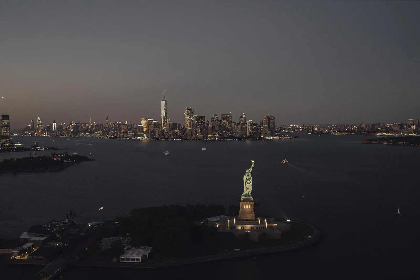 The Statue of Liberty lit at dusk on Liberty Island, with the Manhattan skyline glowing across the harbour behind it