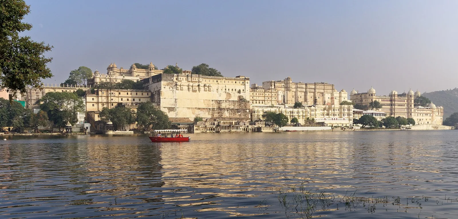 Udaipur City Palace seen across Lake Pichola, with a red passenger boat crossing in front