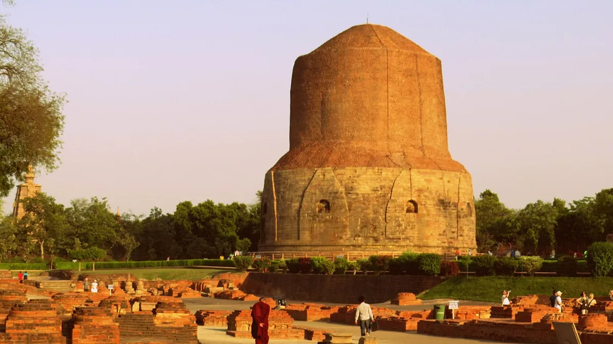 The great brick Dhamek Stupa at Sarnath rising above the excavated monastery ruins, with a Buddhist monk in maroon robes in the foreground